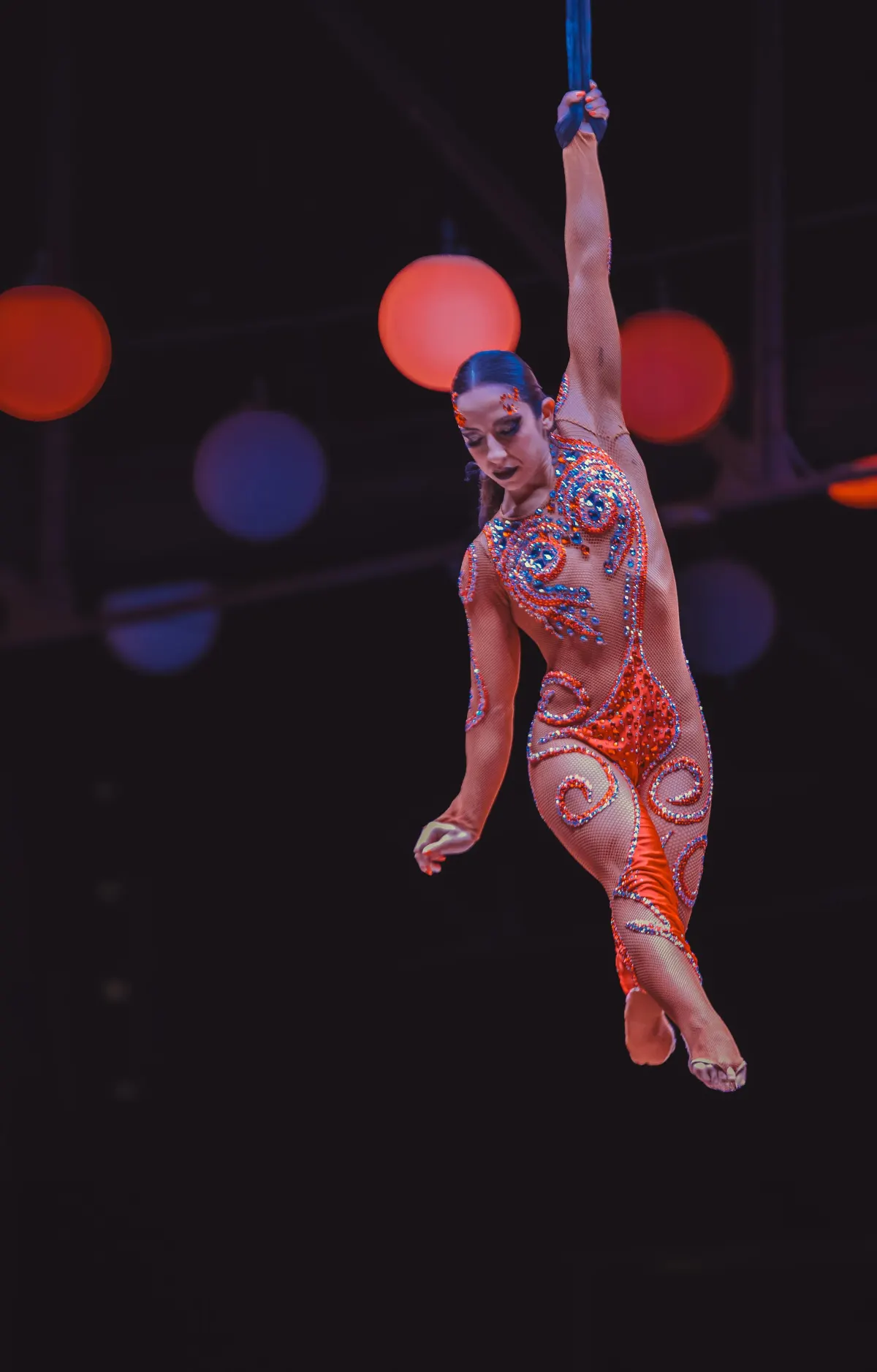 Sara Brigite suspended on an aerial hoop, orange costume and bokeh lights